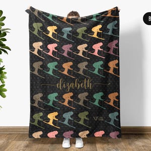 May include: Black blanket featuring a repeating pattern of skiers in various colors. The word "Elizabeth" is written in gold script. The blanket is a cozy home decor item.
