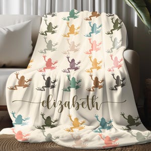 May include: A cream-colored throw blanket featuring a pattern of colorful frogs in various shades. The blanket is draped over a sofa and includes the name "Elizabeth" in a cursive font. The frogs are in shades of green, pink, yellow, and brown.