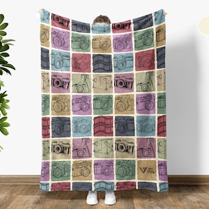 May include: A cream white fleece blanket with a repeating pattern of vintage cameras in various colors. The cameras are drawn in black ink and are set against a background of colorful squares.