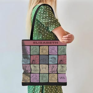 May include: A black tote bag with a grid pattern of coffee-related illustrations in pastel colors. The word "ELIZABETH" is printed in a bold font across the top. The bag has black straps and is being carried by a person wearing a green polka dot dress.