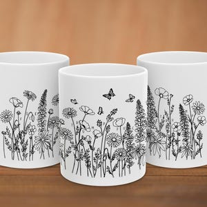 Könnte beinhalten: Drei weiße Keramikbecher mit einer schwarzen Strichzeichnung von Wildblumen und Schmetterlingen. Das Design enthält den Text FUCK TRUMP, der in das florale Muster integriert ist. Jeder Becher hat einen Henkel an der Seite und eine glatte Oberfläche.