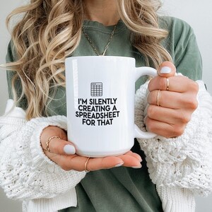 May include: White ceramic mug with the text "I'M SILENTLY CREATING A SPREADSHEET FOR THAT" in black, along with a graphic of a spreadsheet. The mug has a large handle and is held by a person wearing a green shirt and a white sweater.
