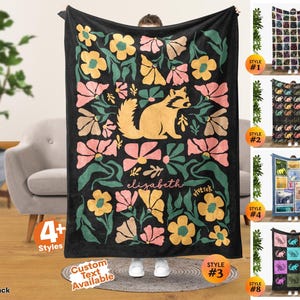 May include: A black rectangular throw blanket featuring a central illustration of a tan raccoon surrounded by stylized pink, yellow, and green flowers. The name elizabeth is printed in a cursive white font below the raccoon, with the text vivtrek nearby.