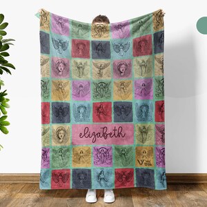 May include: A rectangular fleece blanket featuring a grid pattern of various angel illustrations in shades of pink, blue, yellow, and green. The center of the blanket displays the name elizabeth in a dark script font, with a small logo in the corner.