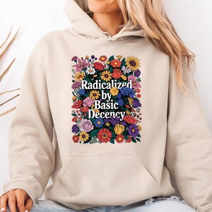 Pode incluir: Um moletom bege com um design floral. O design apresenta uma variedade de flores coloridas em torno do texto "Radicalized by Basic Decency". O moletom tem capuz e um bolso frontal.