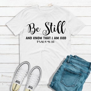 May include: White t-shirt with black text that reads "Be Still and Know That I Am God Psalm 46:10".
