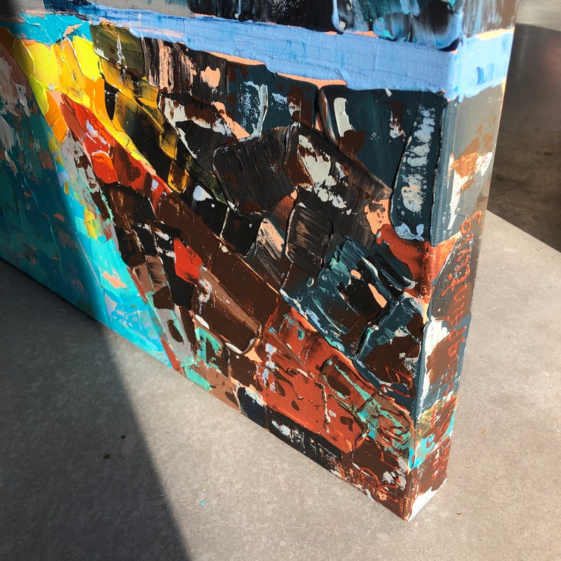 May include: Close-up of the side of a canvas painting with abstract brushstrokes in shades of blue, brown, orange, and yellow. The edges of the canvas are visible.