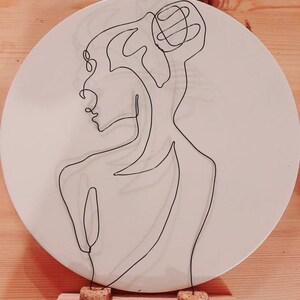 Könnte beinhalten: Ein weißer Keramikteller mit einer schwarzen Draht-Skulptur eines Personenprofils. Die Skulptur ist auf einem Holzsockel mit Korkstopfen befestigt.