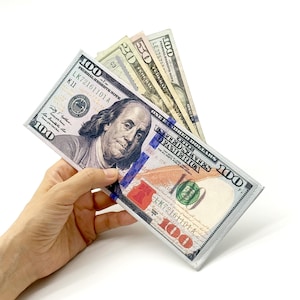 May include: A hand holding a fan of United States currency, including one hundred, fifty, and twenty dollar bills. The bills are displayed against a white background, with the portrait of Benjamin Franklin visible on the $100 bill.