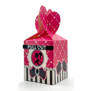 May include: Pink and black favor box with a silhouette of a woman with a crown and the text "PULL OUT" on the front.
