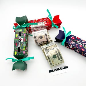 May include: Christmas-themed paper crackers in red, green, and blue, tied with green ribbons. The crackers have festive patterns and the text "PULL OUT". Several crackers are open, revealing US dollar bills.