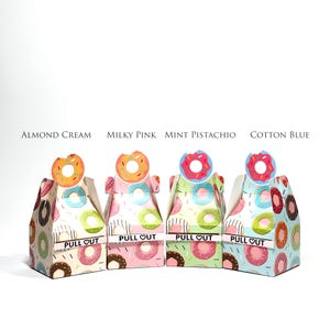May include: Four colorful donut-themed gift boxes. Each box has a different color scheme and a donut-shaped topper. The boxes are labeled "Almond Cream", "Milky Pink", "Mint Pistachio", and "Cotton Blue". The text "PULL OUT" is printed on each box.