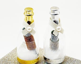 May include: Two clear glass champagne bottles with gold and silver lids. Each bottle contains a rolled up dollar bill with a rhinestone band and a white bow with a pearl. The bottles are filled with white faux fur.