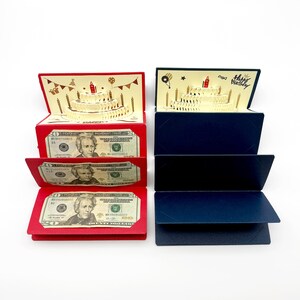 May include: Two multi-tiered pop-up birthday cards, one red and one dark blue. Each card features a three-dimensional paper cake with a red candle. The red card contains three twenty-dollar bills in slots, while the blue card is empty.