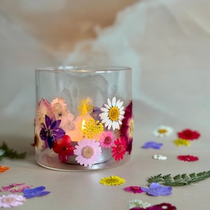 May include: A clear glass candle holder with a tealight candle inside. The holder is decorated with pressed flowers in shades of pink, purple, white, and yellow.