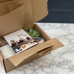 May include: A brown cardboard box with the lid open, revealing a succulent plant kit. The kit includes a succulent plant, soil, decorative stones, and instructions. The box also includes a thank you card.