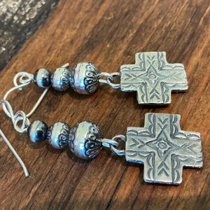May include: Silver earrings with a cross design. Each earring features a decorative cross pendant and a cluster of silver beads. The earrings are displayed on a wooden surface.