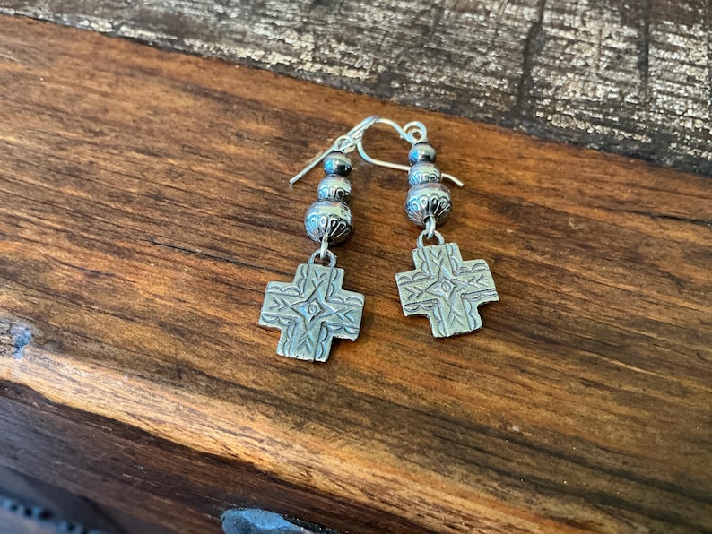 May include: A pair of silver earrings with a cross design. Each earring features a cross pendant with etched details, suspended from a beaded section and a hook. The earrings are displayed on a wooden surface.