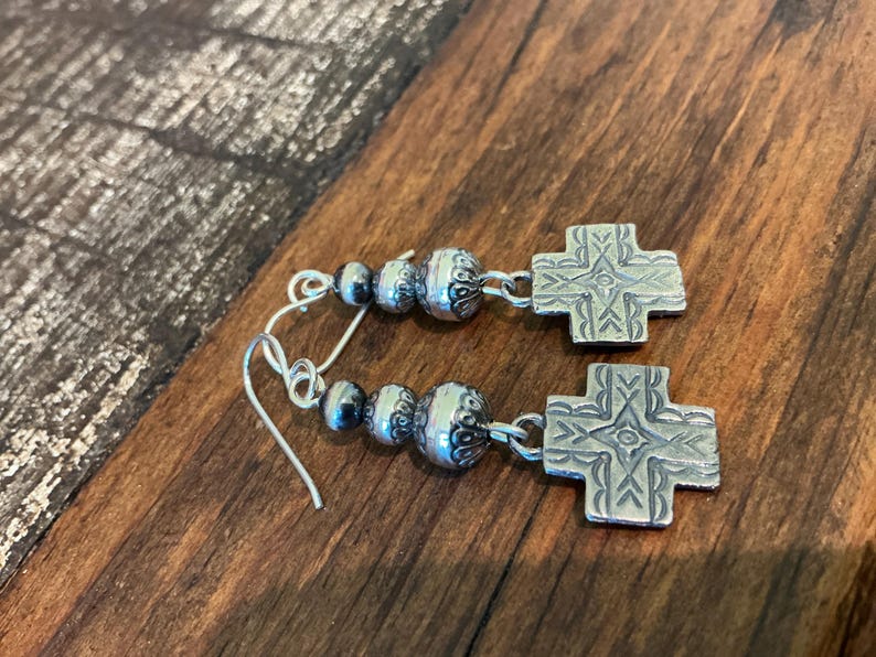 May include: Silver earrings with a cross design. Each earring features a decorative cross pendant and a cluster of silver beads. The earrings are displayed on a wooden surface.