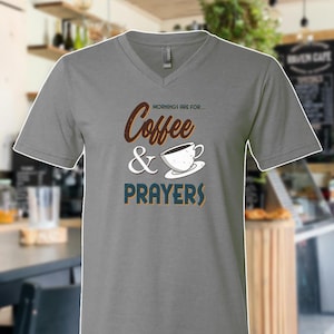 May include: A gray V-neck t-shirt with the words "Mornings are for... Coffee & Prayers" in brown, white, and blue. The shirt features a coffee cup graphic. The background is a blurred cafe setting.