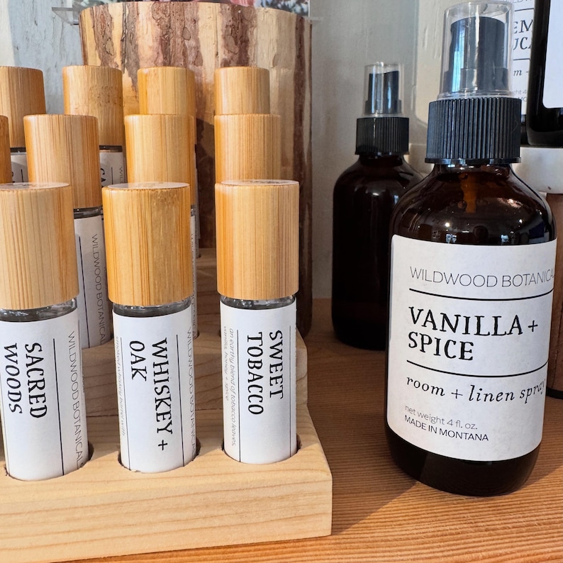 Puede incluir: Una exhibici&oacute;n de aerosoles para habitaciones y ropa de cama y roll-ons de aceites esenciales de Wildwood Botanicals. El spray Vanilla + Spice est&aacute; en una botella de vidrio marr&oacute;n. Los roll-ons incluyen Sacred Woods, Whiskey + Oak y Sweet Tobacco.