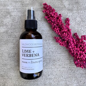 Puede incluir: Una botella de vidrio marr&oacute;n de spray para habitaciones y ropa de cama Wildwood Botanica Lime + Verbena, con un pulverizador negro y una etiqueta blanca. La etiqueta dice "net weight 4 fl oz. MADE IN MONTANA". Un vibrante tallo floral rosa est&aacute; al lado de la botella.