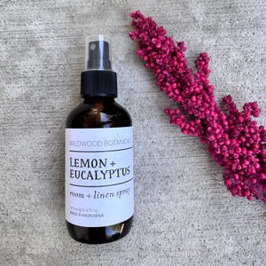 Puede incluir: Una botella de vidrio marr&oacute;n de spray para habitaciones y ropa de cama Lemon + Eucalyptus de Wildwood Botanicals. La etiqueta es blanca con texto negro. Un tallo floral rosa est&aacute; al lado de la botella. La botella es de 118 ml.