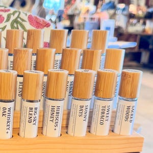 Puede incluir: Una exhibici&oacute;n de frascos roll-on de aceites esenciales con tapas de madera. Las etiquetas indican "Whiskey + Oak", "Big Sky Blend", "Hibiscus + Honey", "Sunny Days", "Sweet Tobacco" y "Sacred Woods". Los frascos est&aacute;n en un expositor de madera.