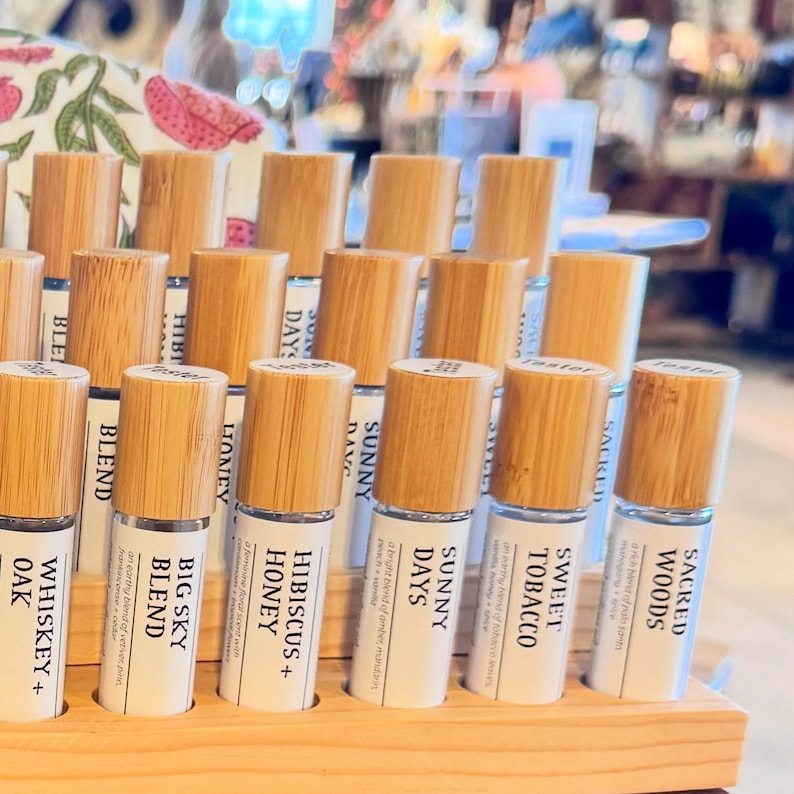 Puede incluir: Una exhibici&oacute;n de frascos roll-on de aceites esenciales con tapas de madera. Las etiquetas indican "Whiskey + Oak", "Big Sky Blend", "Hibiscus + Honey", "Sunny Days", "Sweet Tobacco" y "Sacred Woods". Los frascos est&aacute;n en un expositor de madera.