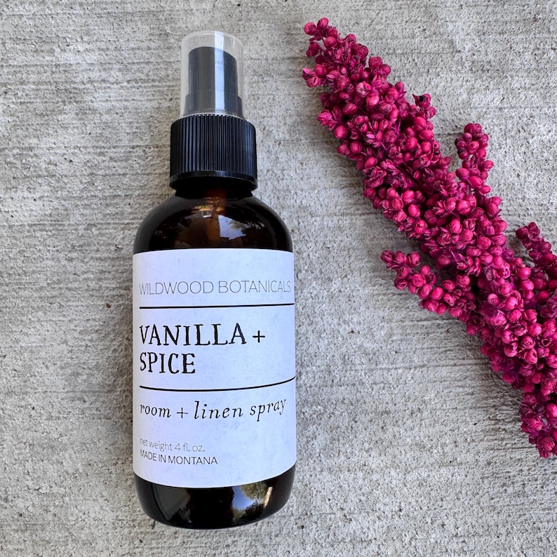 Puede incluir: Una botella de vidrio marr&oacute;n de spray para habitaciones y ropa de cama Wildwood Botanicals Vanilla + Spice. La etiqueta indica un peso neto de 118 ml. Una ramita de flores magenta est&aacute; al lado de la botella. Hecho en Montana.