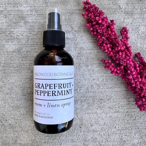 Puede incluir: Una botella de spray de vidrio &aacute;mbar oscuro con una tapa negra y una boquilla de spray transparente. La etiqueta dice "WILDWOOD BOTANICALS GRAPEFRUIT + PEPPERMINT room + linen spray, peso neto 118 ml, MADE IN MONTANA." Un tallo de flor magenta est&aacute; a la derecha.