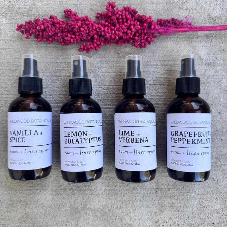 Puede incluir: Cuatro botellas de spray de vidrio marr&oacute;n con atomizadores negros, cada una etiquetada con un aroma diferente: Vainilla + Especias, Lim&oacute;n + Eucalipto, Lima + Verbena y Pomelo Menta. Un tallo floral rosa est&aacute; encima de las botellas.