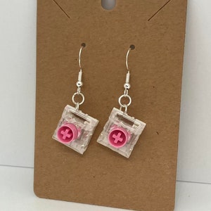 Könnte beinhalten: Ohrringe mit transparenten, quadratischen Anhängern, die jeweils ein rosa Pluszeichen aufweisen. Die Ohrringe haben silberfarbene Haken und werden vor einem braunen Hintergrund präsentiert.