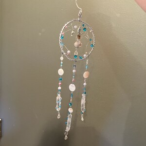 May include: A handcrafted dreamcatcher with a silver wire frame. The frame is adorned with pink, blue, and white beads, along with clear quartz crystals and shell accents. A small glass bottle and a dolphin charm are suspended within the frame.