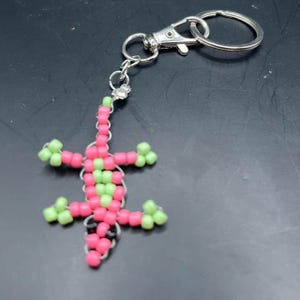 May include: A beaded keychain featuring a lizard design. The lizard is crafted with pink and green beads, and has a silver-colored metal clasp and key ring. The keychain is a handmade accessory.