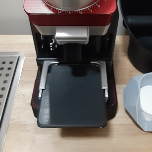 Puede incluir: Un molinillo de café rojo y negro con una tapa de madera y un recipiente de recogida negro. El molinillo tiene un temporizador digital y una bandeja negra para recoger el café molido.