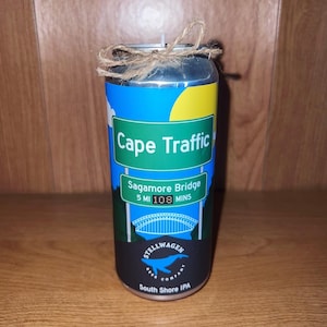 May include: A silver can candle with a blue and yellow design. The can has a green sign that says "Cape Traffic" and a smaller sign that says "Sagamore Bridge 5 MI 108 MINS". The can also has a black and white logo that says "Stellwagen Beer Company" and "South Shore IPA".