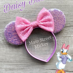 May include: Purple and pink sequined Minnie Mouse ears with a pink bow and a purple headband. The headband has the text "Main Street Mickey" written on it. Daisy Duck is pictured in the bottom right corner of the image.