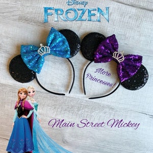 May include: Two pairs of black sequin Minnie Mouse ears with a headband. One pair has a blue bow with a silver crown and the other has a purple bow with a silver crown. The text "More Princesses!" is written on the headband. The text "Main Street Mickey" is written below the ears.