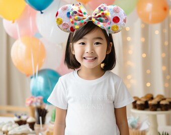 May include: A young girl wearing a white shirt with the text "Main Street Mickey" on it. She is wearing a colorful polka dot headband with a bow. The headband is shaped like Mickey Mouse ears. The girl is smiling and looking at the camera. There are balloons and cupcakes in the background.
