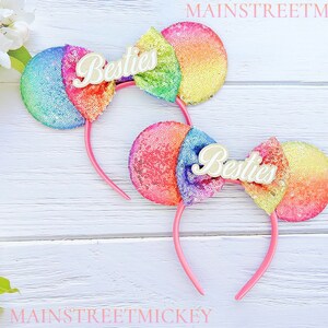 Puede incluir: Dos diademas con lentejuelas con orejas de colores arcoíris y diademas rosas. Cada diadema tiene un lazo color crema con la palabra "Besties" en escritura cursiva. Las diademas están sobre una superficie de madera blanca.