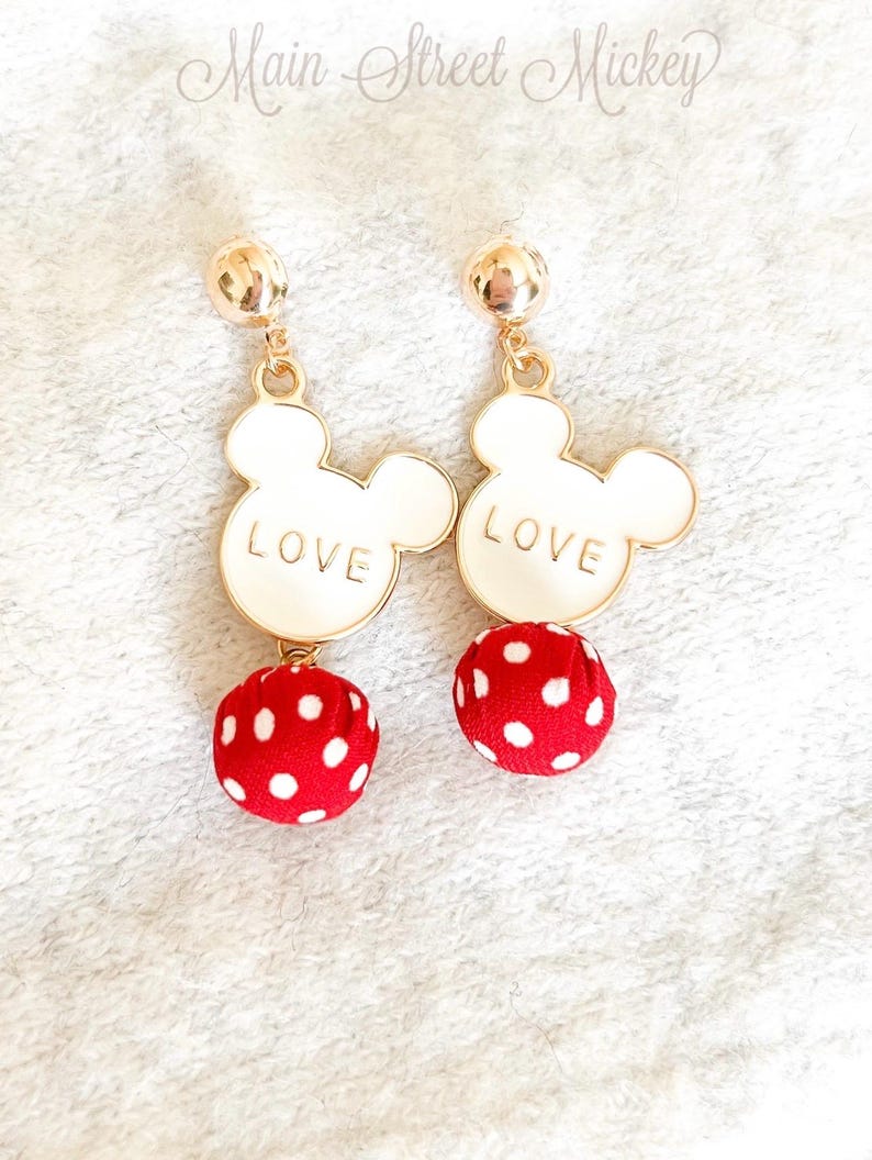 Może przedstawiać: Para złotych kolczyk&oacute;w. Każdy kolczyk ma białą głowę Myszki Miki z napisem "LOVE" i czerwoną kulkę z białymi kropkami. Kolczyki mają zapięcie na sztyft.