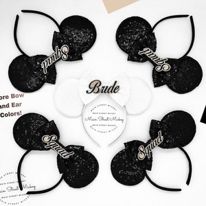 Puede incluir: Cuatro orejas de Minnie Mouse de lentejuelas negras con letras doradas brillantes. Las orejas están etiquetadas como "Bride", "Squad" y "Squad".