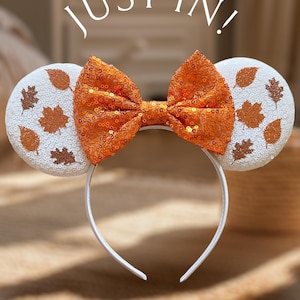 Puede incluir: Orejas de ratón blancas con un lazo de lentejuelas naranja y detalles de hojas marrones. Las orejas están decoradas con lentejuelas blancas y hojas marrones. La diadema es blanca. "Just In!" está escrito en blanco sobre un fondo borroso.