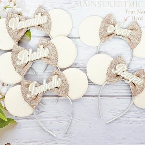May include: Five pairs of white mouse ears with gold glitter bows and personalized names. The names are Veronika, Blaike, Isabelle, Noor, and Reagan.