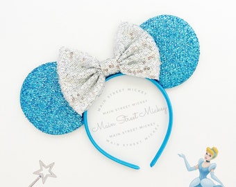 May include: Light blue and silver glitter Minnie Mouse ears with a blue headband that says "Main Street Mickey" on it. Cinderella is in the background.