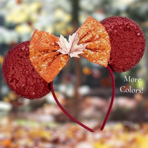 May include: Red and orange sequined Minnie Mouse ears with a brown felt bow and a brown felt maple leaf. The headband is red.