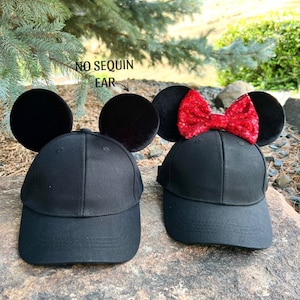 May include: Two black baseball caps with black Mickey Mouse ears. One cap has a red sequin bow on the ear. The text "NO SEQUIN EAR" is written on the image.