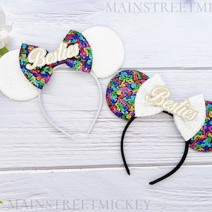 Puede incluir: Dos diademas con orejas de lentejuelas blancas y lazos de lentejuelas de colores. Los lazos tienen la palabra "Besties" en cursiva. Una diadema es blanca y la otra negra. El fondo es una superficie de madera blanca.