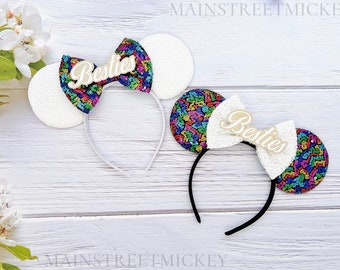 Könnte beinhalten: Zwei Haarbänder mit weißen Paillettenohren und bunten Pailletten-Schleifen. Die Schleifen tragen das Wort "Besties" in Schreibschrift. Ein Stirnband ist weiß, das andere schwarz. Der Hintergrund ist eine weiße Holzoberfläche.